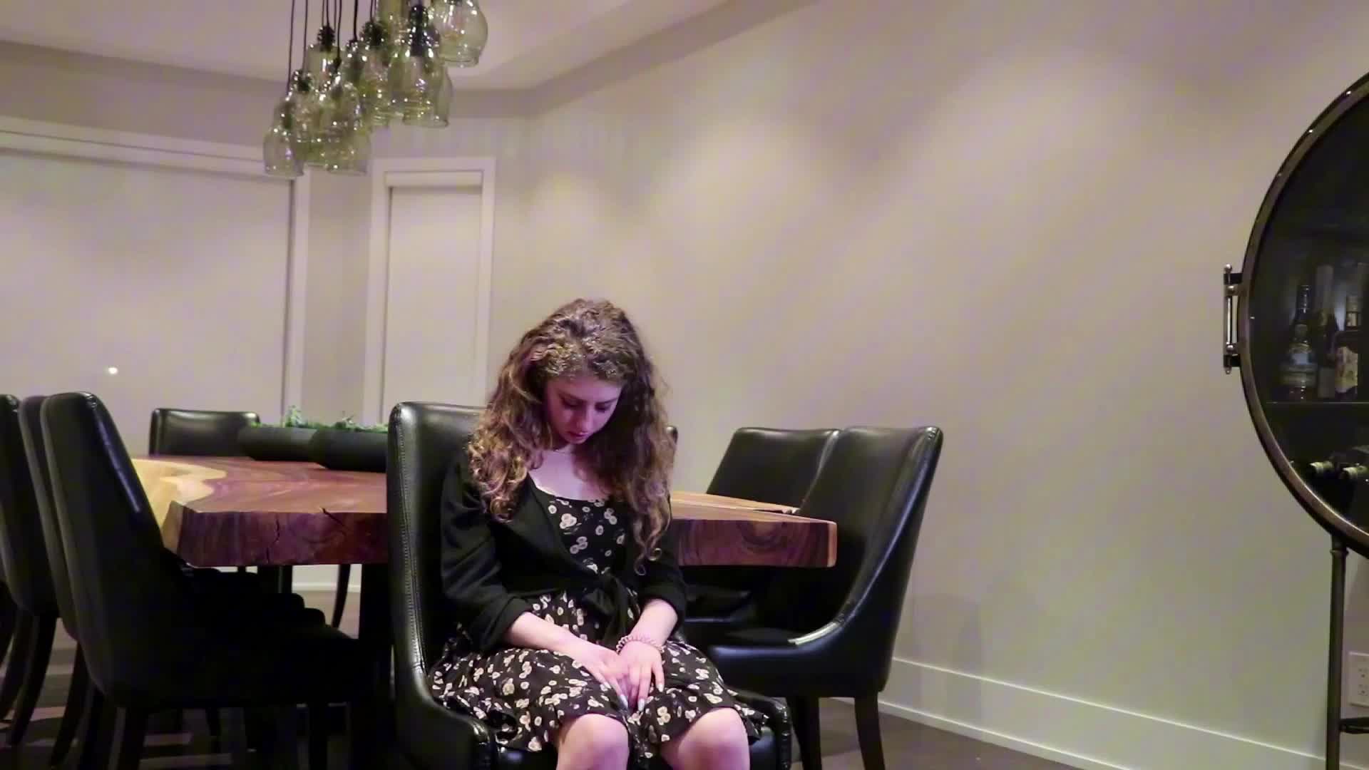 A young woman with curly hair sits on a black chair, wearing a black and white floral dress, with her head bowed and hands resting on her lap, in a modern dining room with a large wooden table and pendant lights.