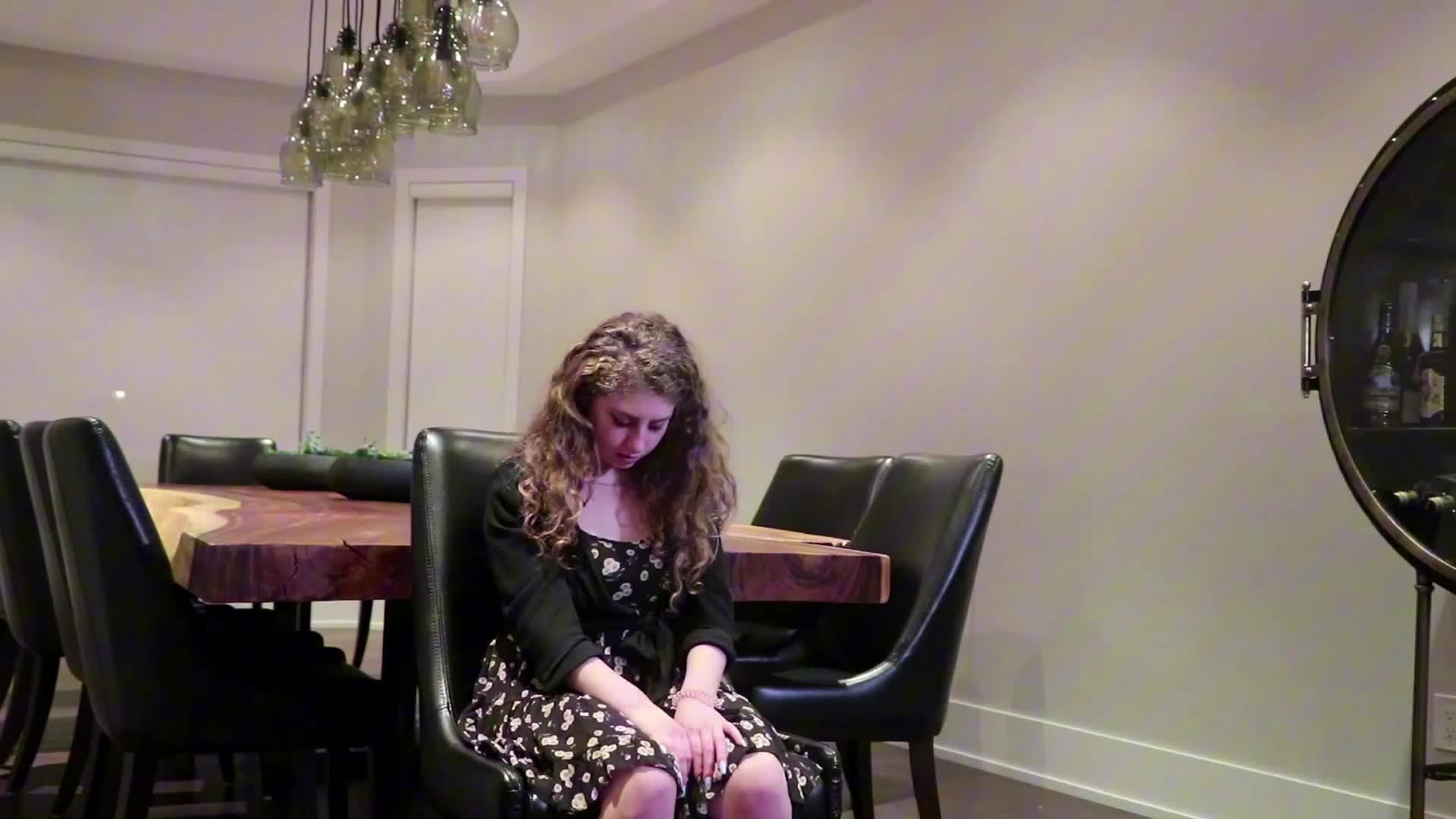 A young woman with curly hair sits on a black chair, wearing a floral dress and a black cardigan, with her head bowed and hands resting on her knees, in a modern dining room with a wooden table and pendant lights.