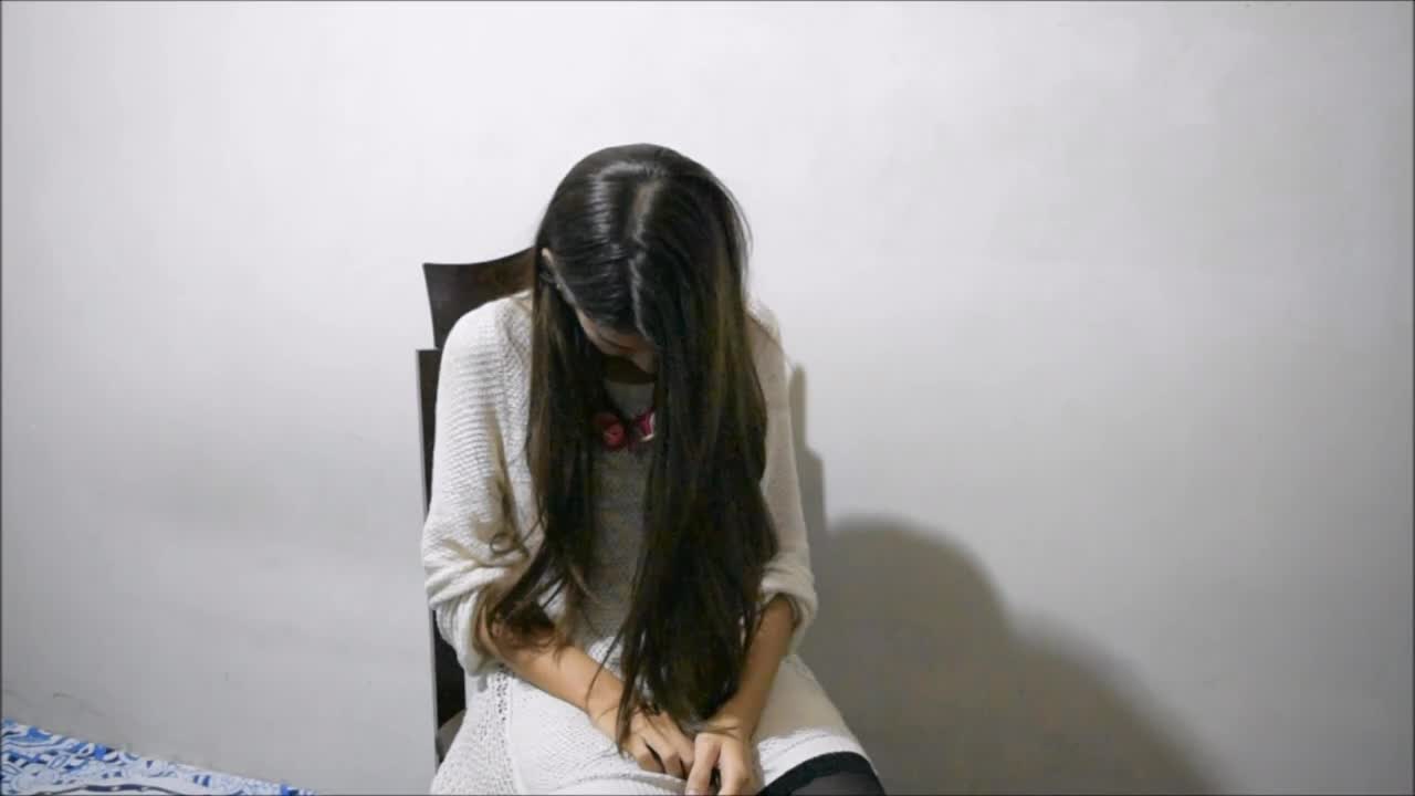 A young woman with long hair is sitting on a chair, looking down with her hands clasped in her lap, against a plain white wall.