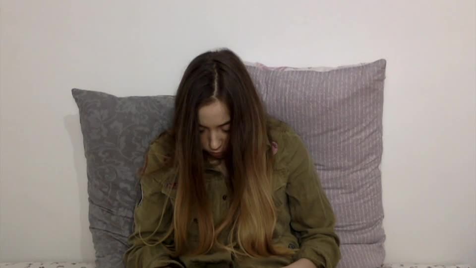 A young woman with long hair is sitting on a bed, looking down at her phone, surrounded by large pillows.