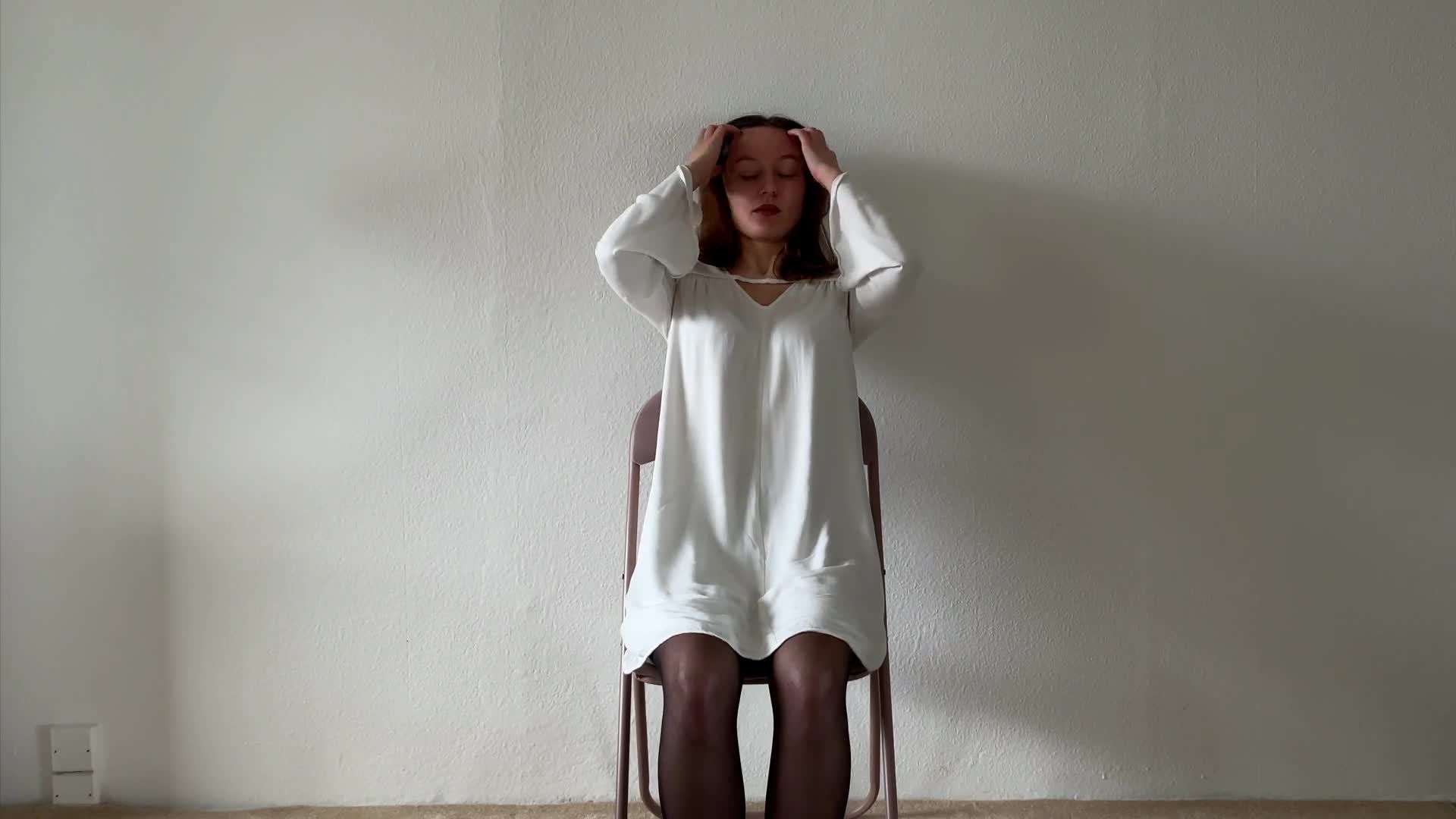 A woman in a white dress sits on a chair with her hands on her head, against a plain white wall.