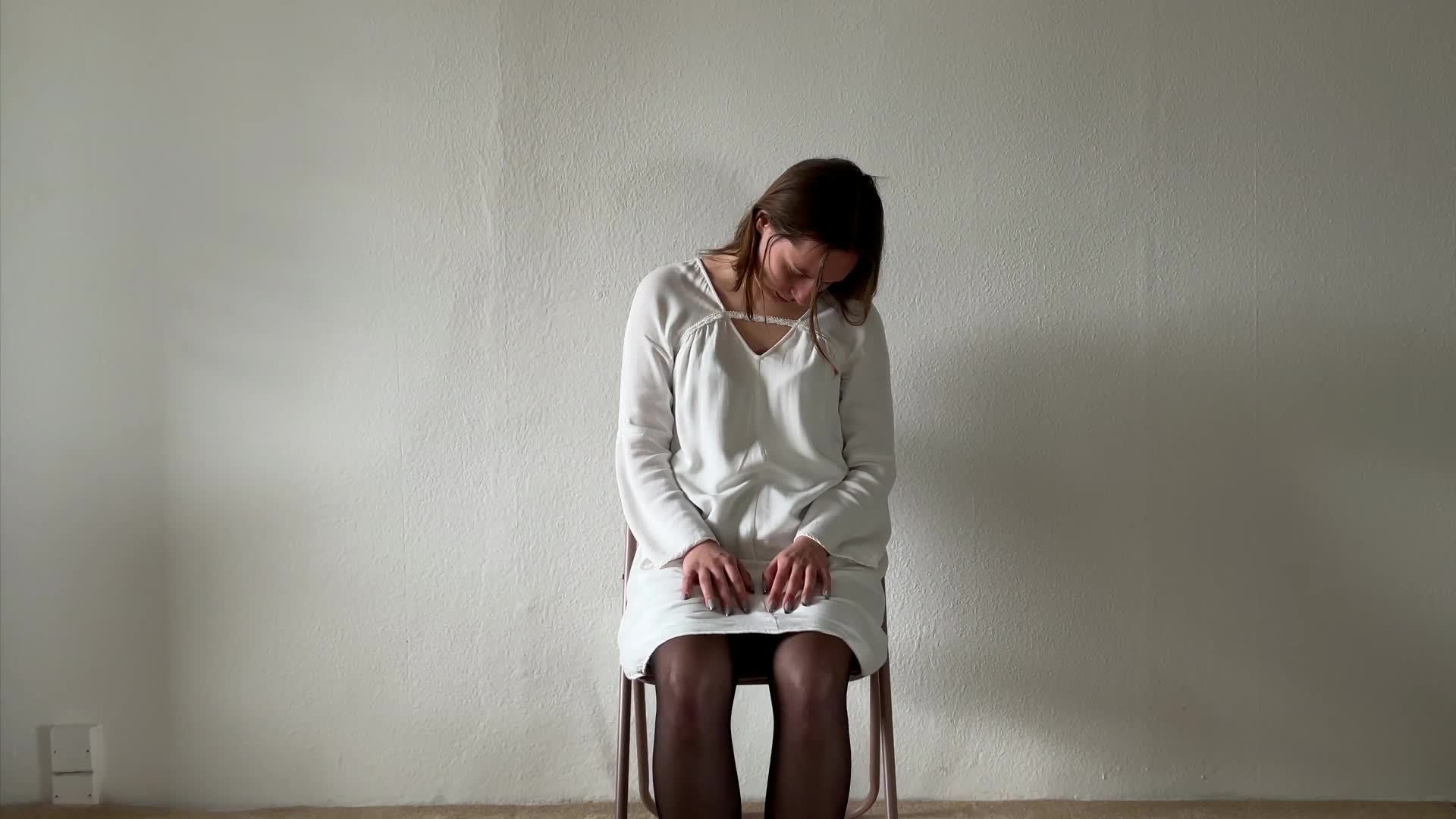 A woman in a white dress sits on a chair with her head bowed and hands resting on her knees, against a plain white wall.