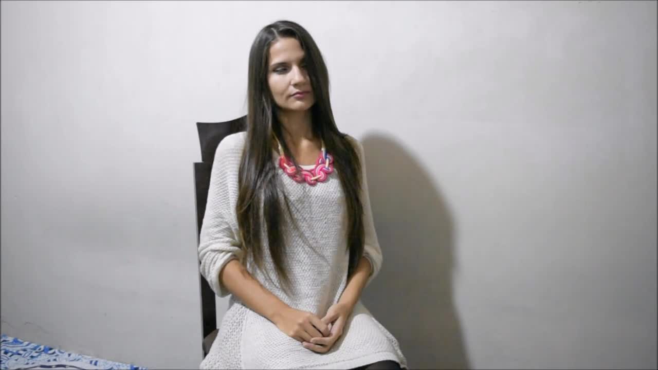 A woman with long hair sits on a chair wearing a white sweater and a colorful necklace against a plain wall.