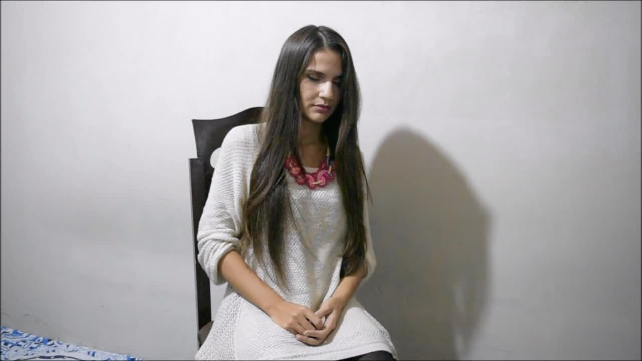 A young woman with long hair sits on a chair, wearing a white sweater and a colorful necklace, against a plain wall.